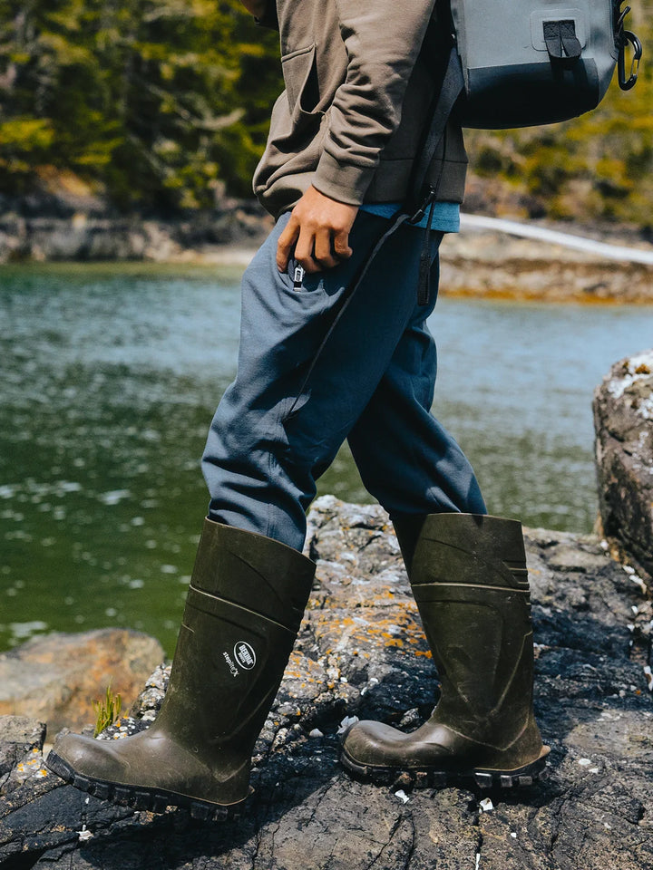 Person wearing green rubber boots standing on rocks near a body of water with trees in the background.